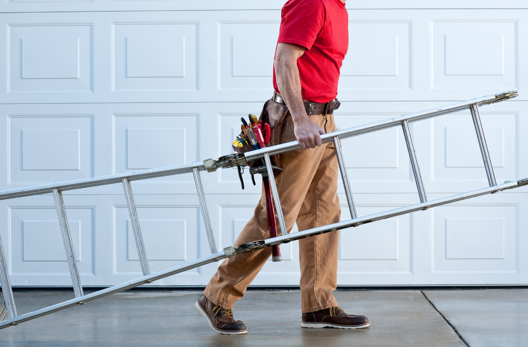 A handyman wearing a red shirt and brown work pants walks in front of a white garage door while carrying an aluminum ladder. He has a tool belt equipped with various tools, including a screwdriver and pliers, signifying home repair and maintenance services.